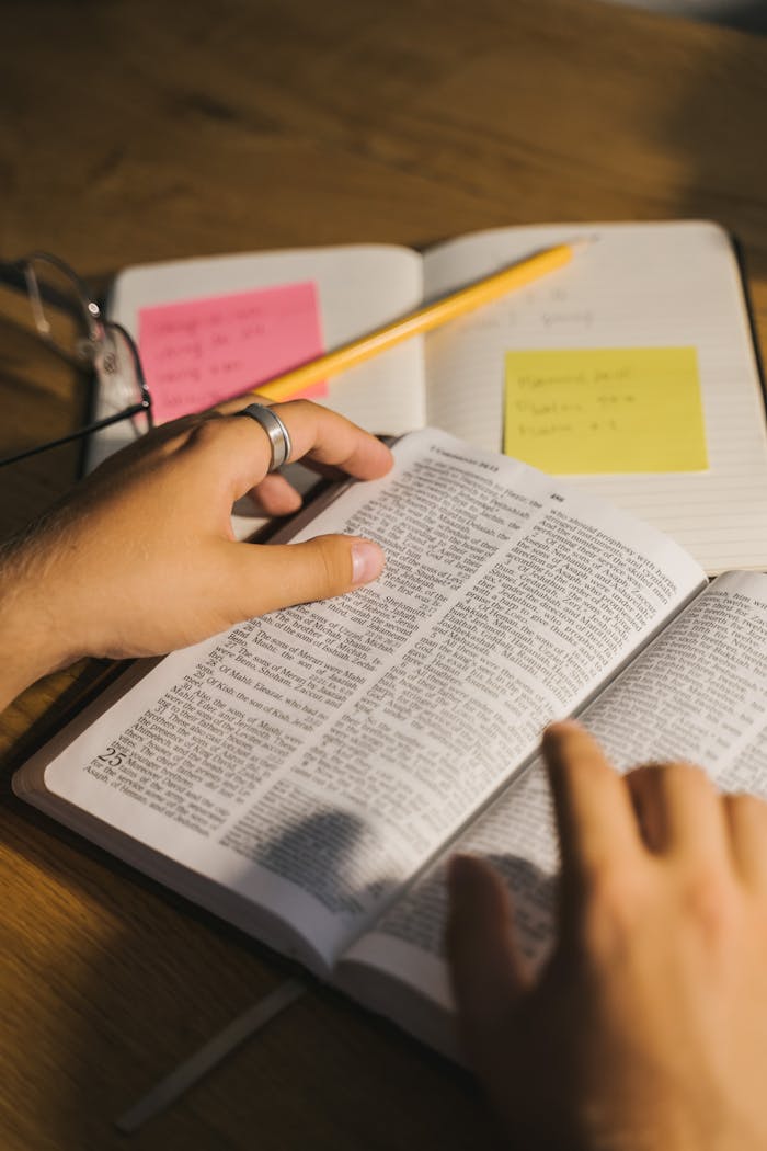 about-us Close-up of hands holding and reading a Bible with study notes and a pencil on a desk.